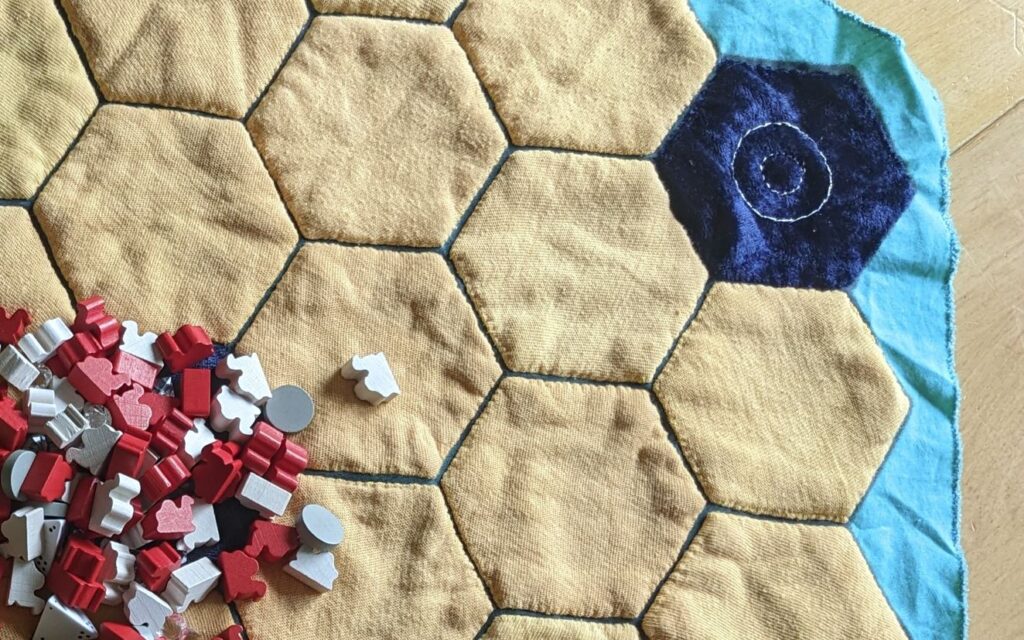 Gelbe Stoff-Sechsecke und ein blaues, aufgenäht auf einen dünnen türkisen Stoff, darauf ein Häufchen Kamelspielsteine